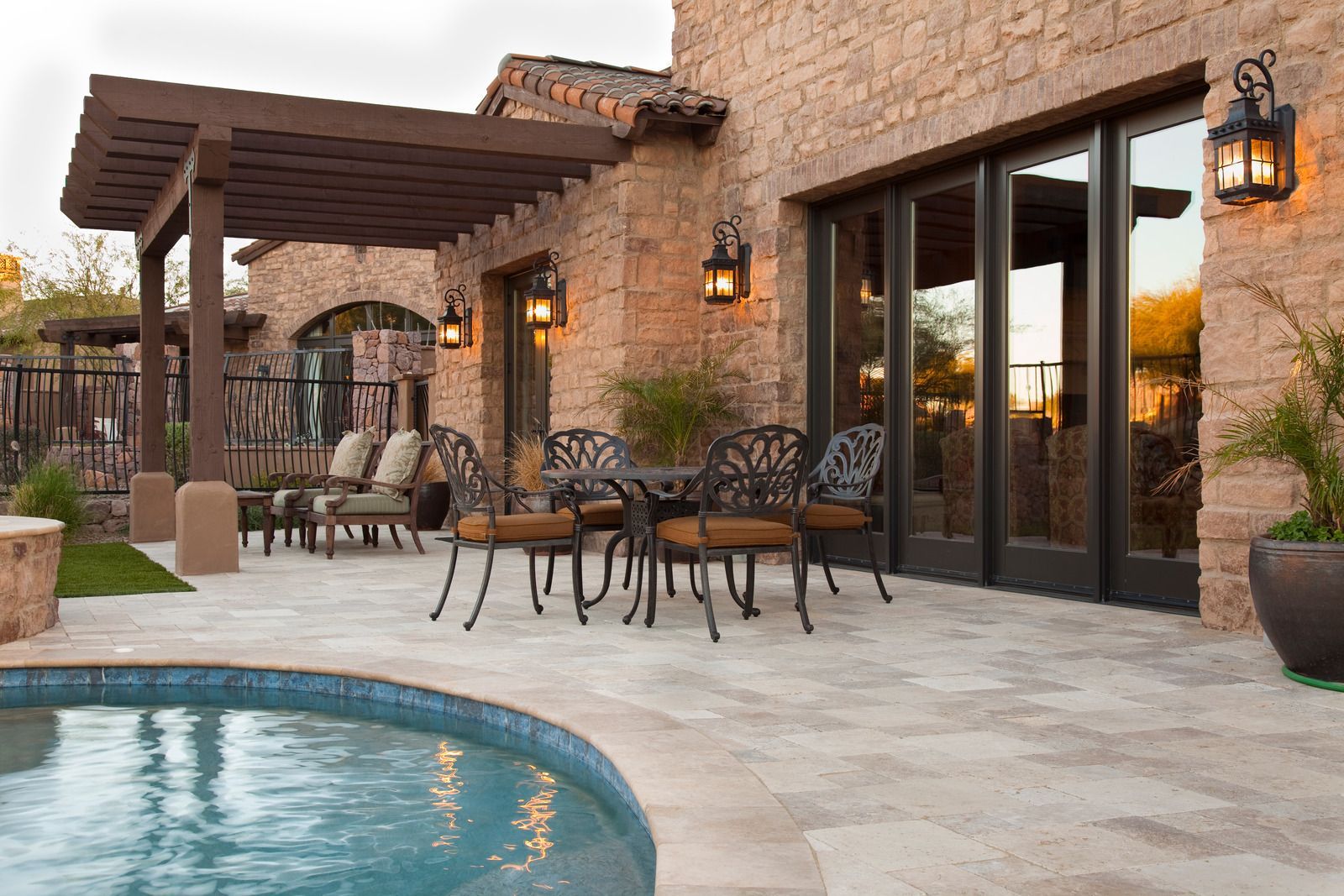 Stone patio with wrought-iron seating beside a pool and a pergola attached to a stucco house at dusk