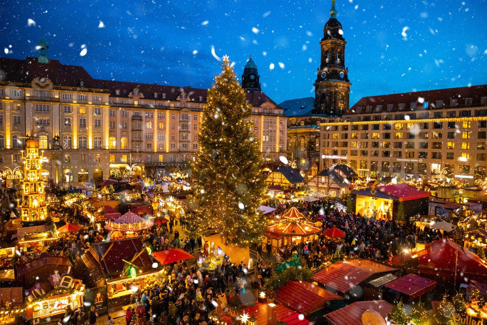 Festive Christmas market at night with glowing stalls, a large decorated tree, and historic buildings under snow.