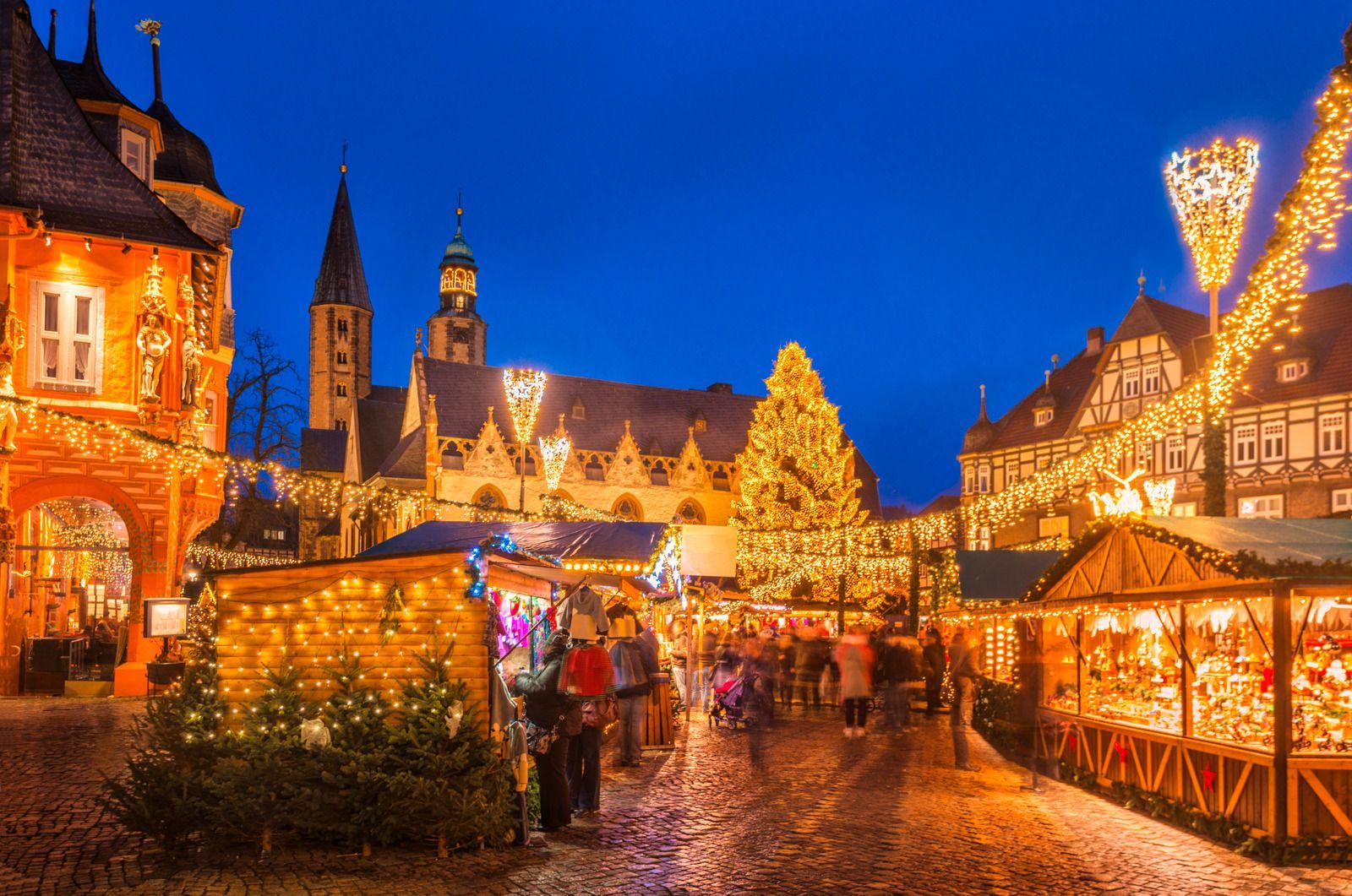 Festive Christmas market at dusk, with glowing lights, crowded stalls, and a cathedral tower in the background