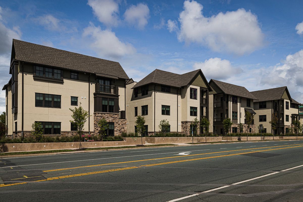 Row of beige apartment buildings with dark roofs along a quiet street under a blue sky