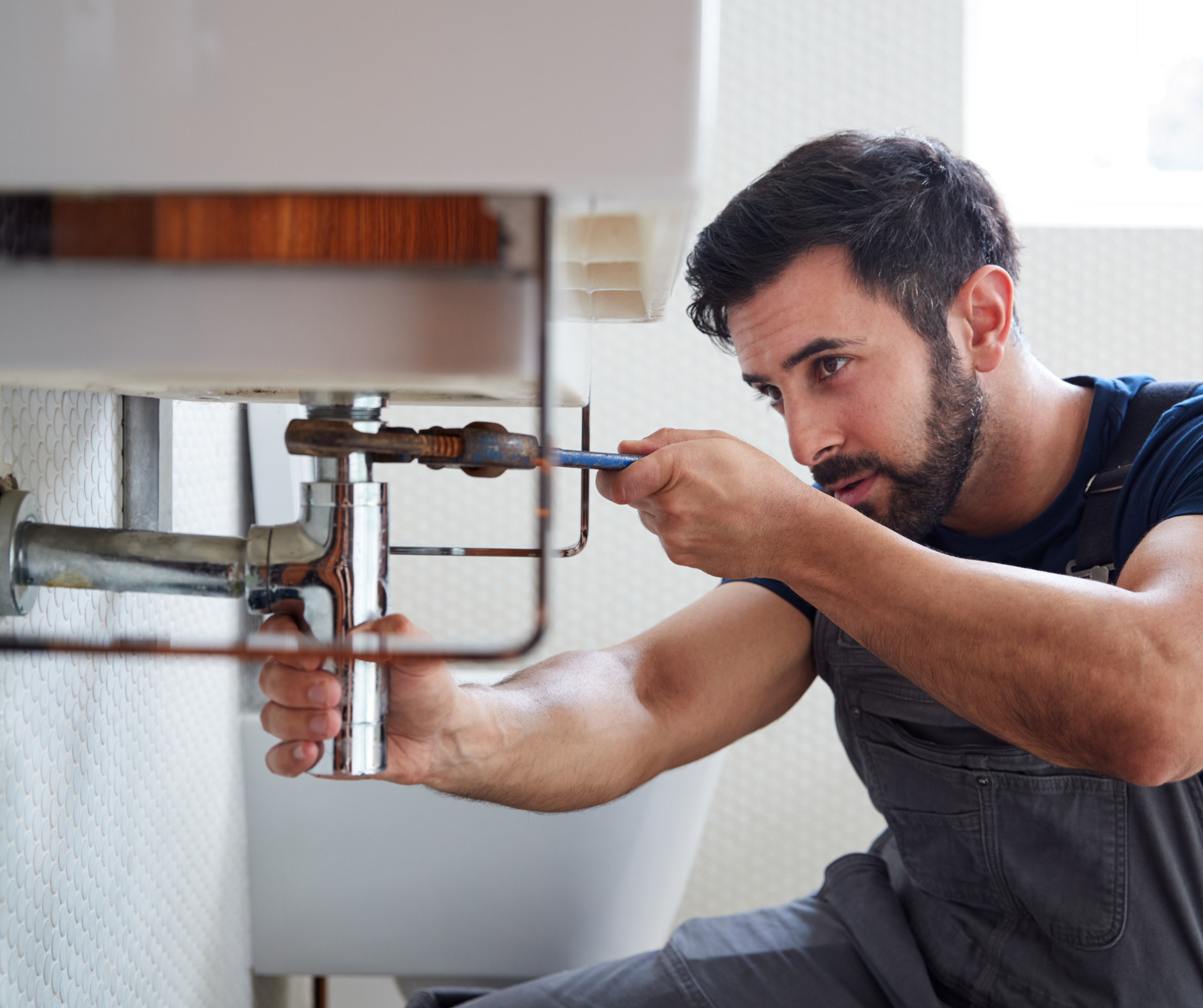 Plumber working under a sink, using a wrench. Indoors, focused expression.