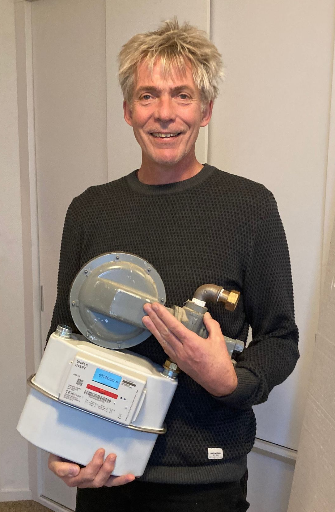 Een man met een gasmeter en bijbehorende onderdelen staat binnen te glimlachen vanwege gasvrij.