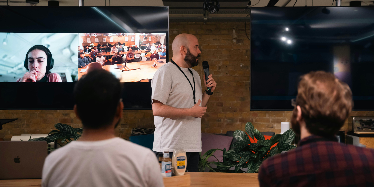 A person speaks into a microphone before an audience, with large screens displaying a video call in a brick-walled room.