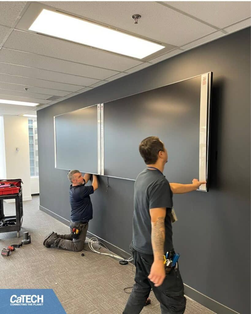 Two men installing a large, silver-framed display on a dark gray wall in an office.