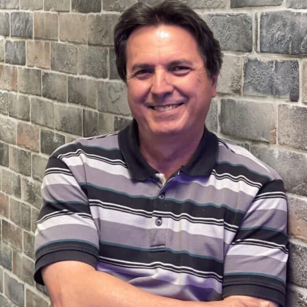 Man with dark hair, smiling, leaning against a brick wall, wearing a striped polo shirt.