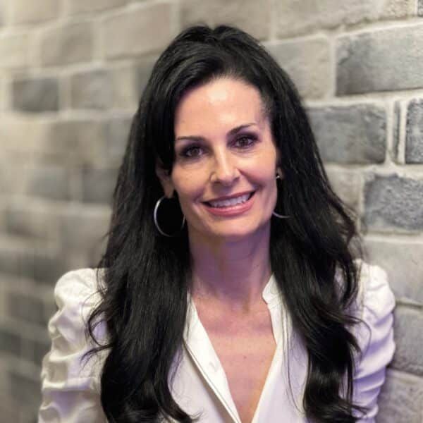 Woman with long dark hair smiling, leaning against a brick wall, wearing a white blouse and large hoop earrings.