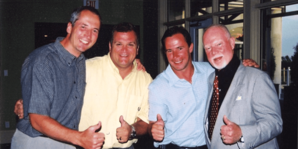 Four men smiling and giving thumbs up in a room with large windows.