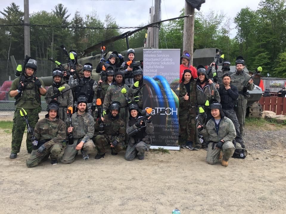 Group of people in paintball gear pose with banner at outdoor paintball field.