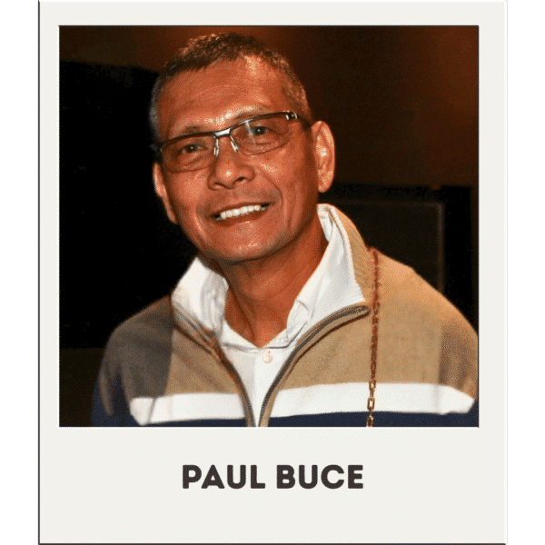 Paul Buce smiles at the camera, wearing glasses, a sweater, and a white collared shirt.