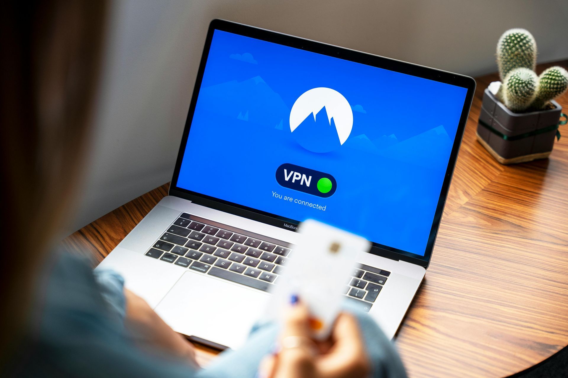 Person using a laptop showing a VPN app, with a phone in hand and a small cactus on the desk.