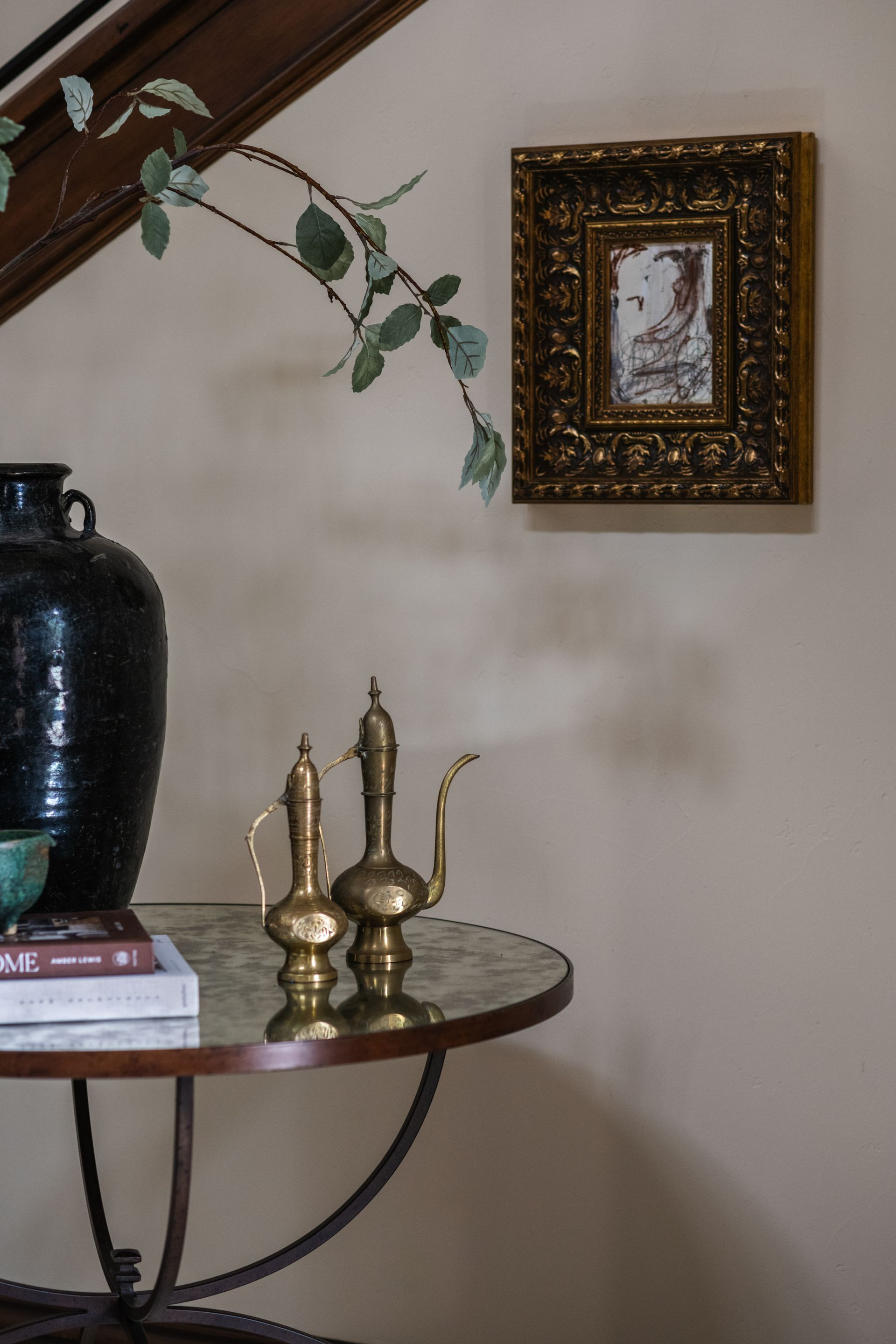 Decorative corner table with black vase, brass candlesticks, books, and framed wall art with a leafy branch above.