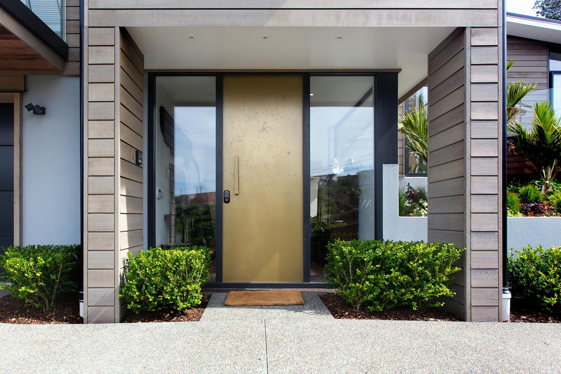 Modern home entrance with gold door, glass sidelights, and trimmed shrubs outside.