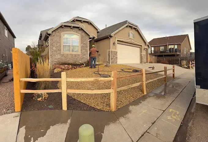 A split-rail fence surrounds a rocky landscape in front of a tan house with a light-colored garage.