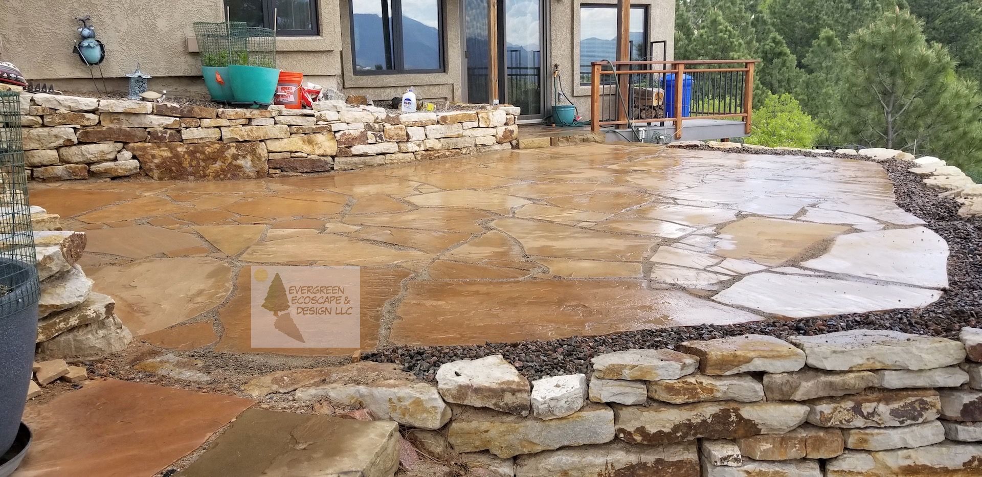 Stone patio with wet tan concrete and stone edging outside a house, with a small railing and potted plant.