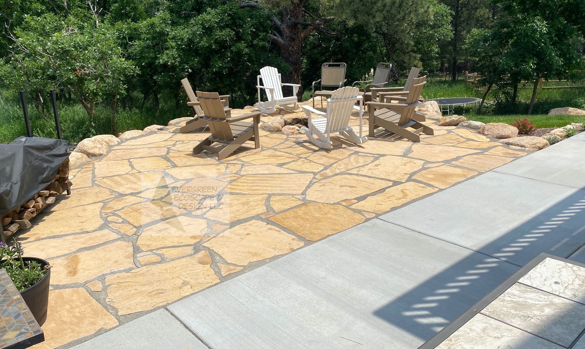Stone patio with Adirondack chairs and a table beside a concrete walkway in a garden setting