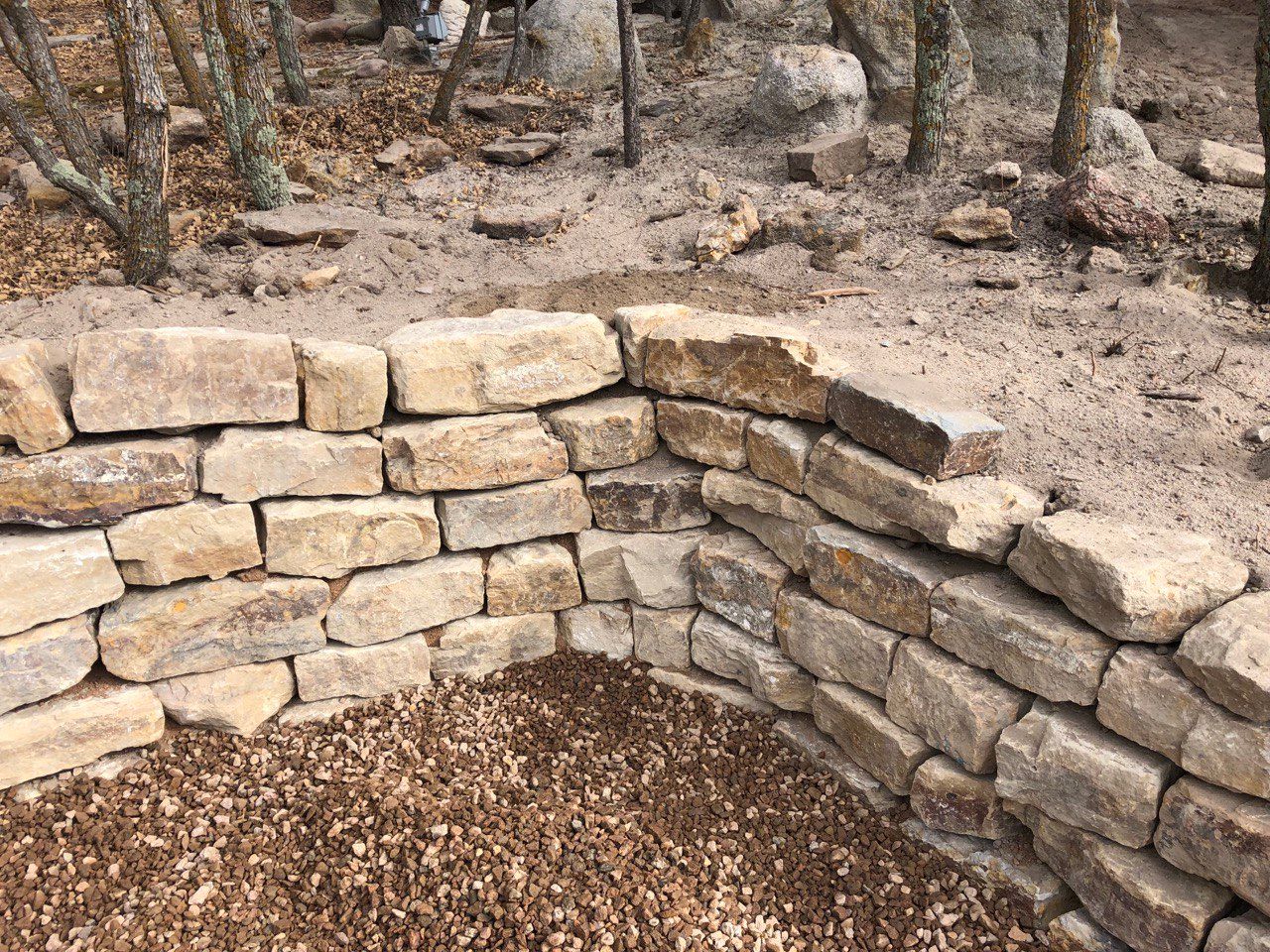Stone retaining wall curving around a leaf-covered outdoor area in a wooded setting