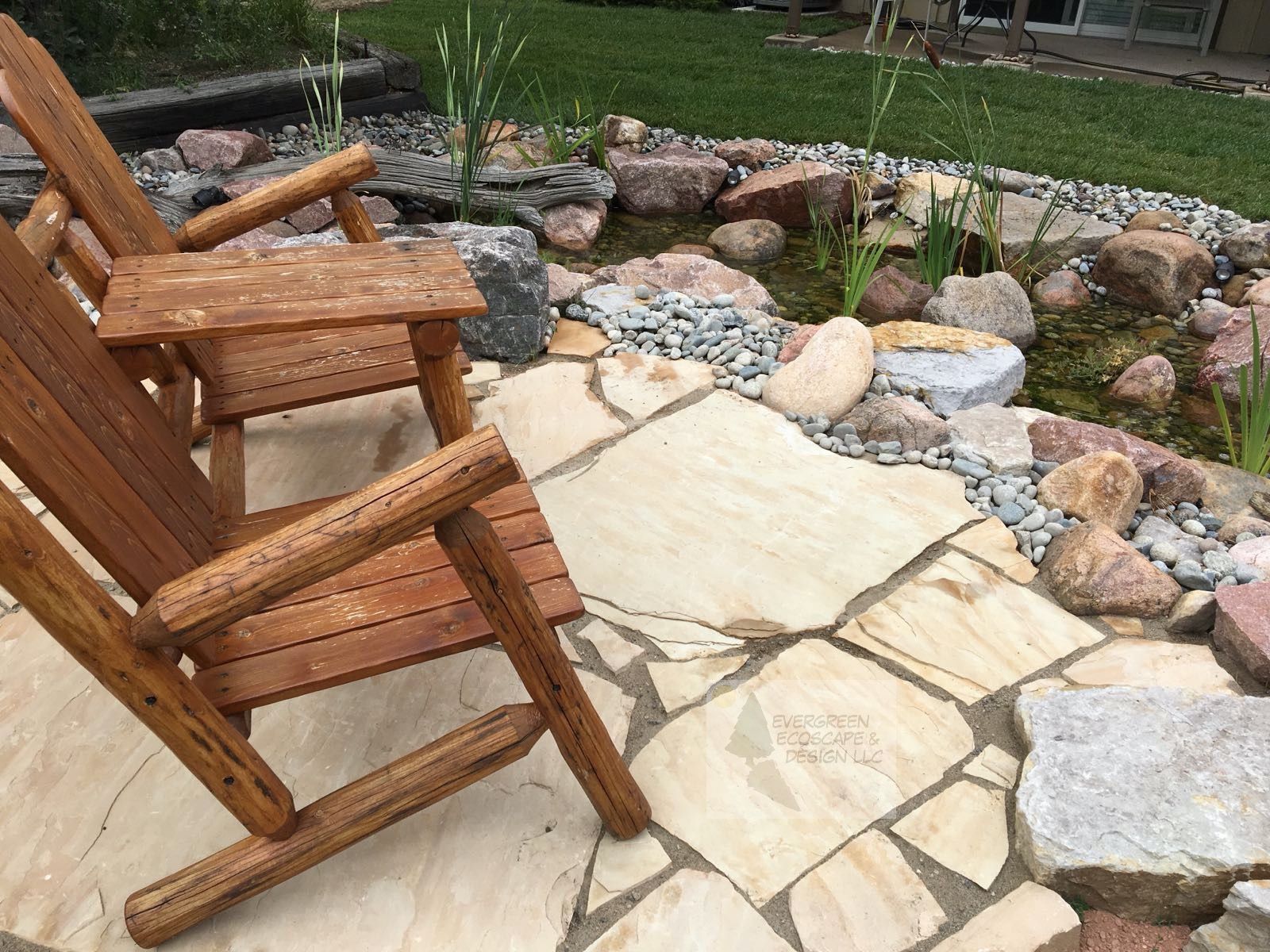 Wooden chairs on a stone patio beside a small rock garden with a stream and stones.
