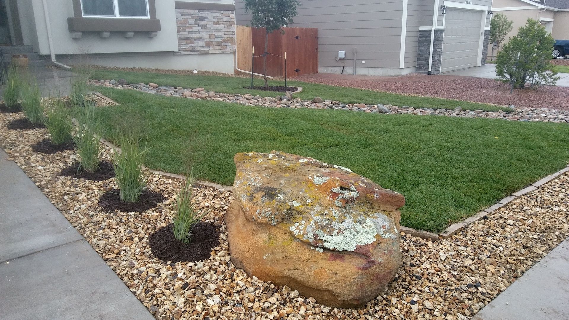 Front yard landscaping with green lawn, rock borders, small shrubs, and a large decorative stone planter.