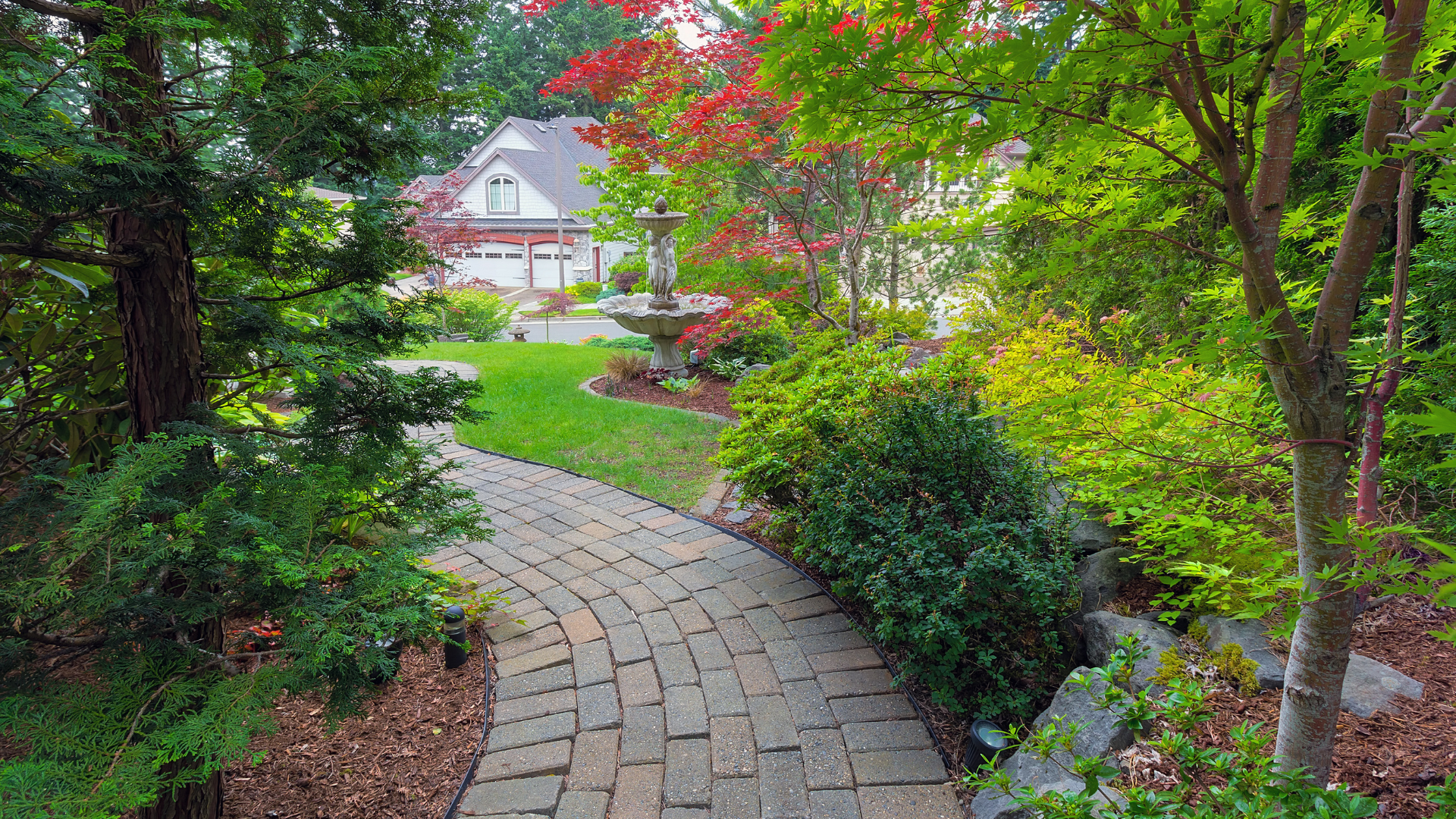 Curved brick garden path leading through lush greenery to a house with a porch and red-leafed tree