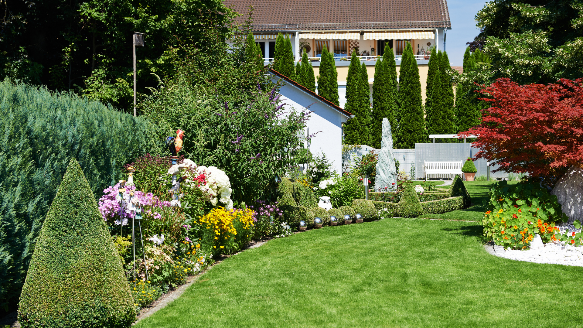 Lush garden with colorful flowers, green lawn, tall shrubs, and a house in the background