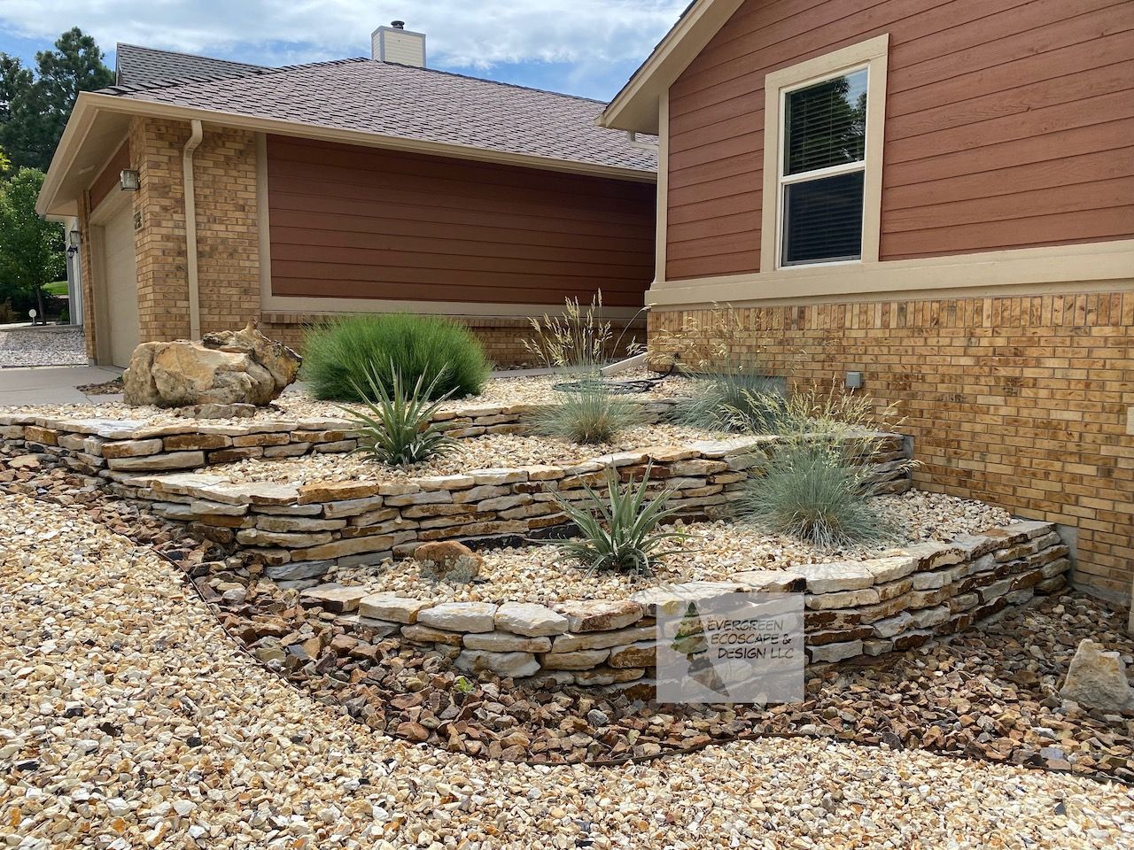 Stone terraced landscaping beside a tan house with gravel and desert plants under a cloudy sky