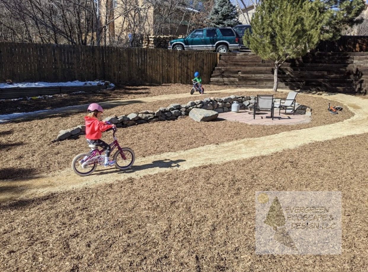 Two children riding bikes on a gravel path in a backyard with a fire pit seating area