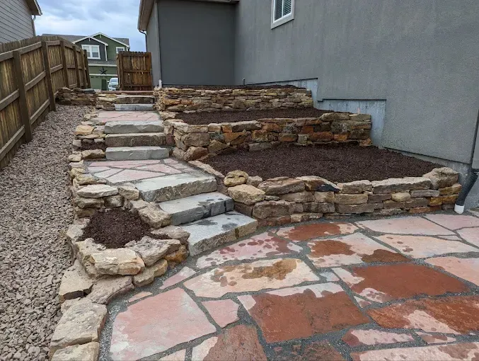 Stone steps and retaining walls with mulch beds leading to a wooden fence. Patio in the foreground.
