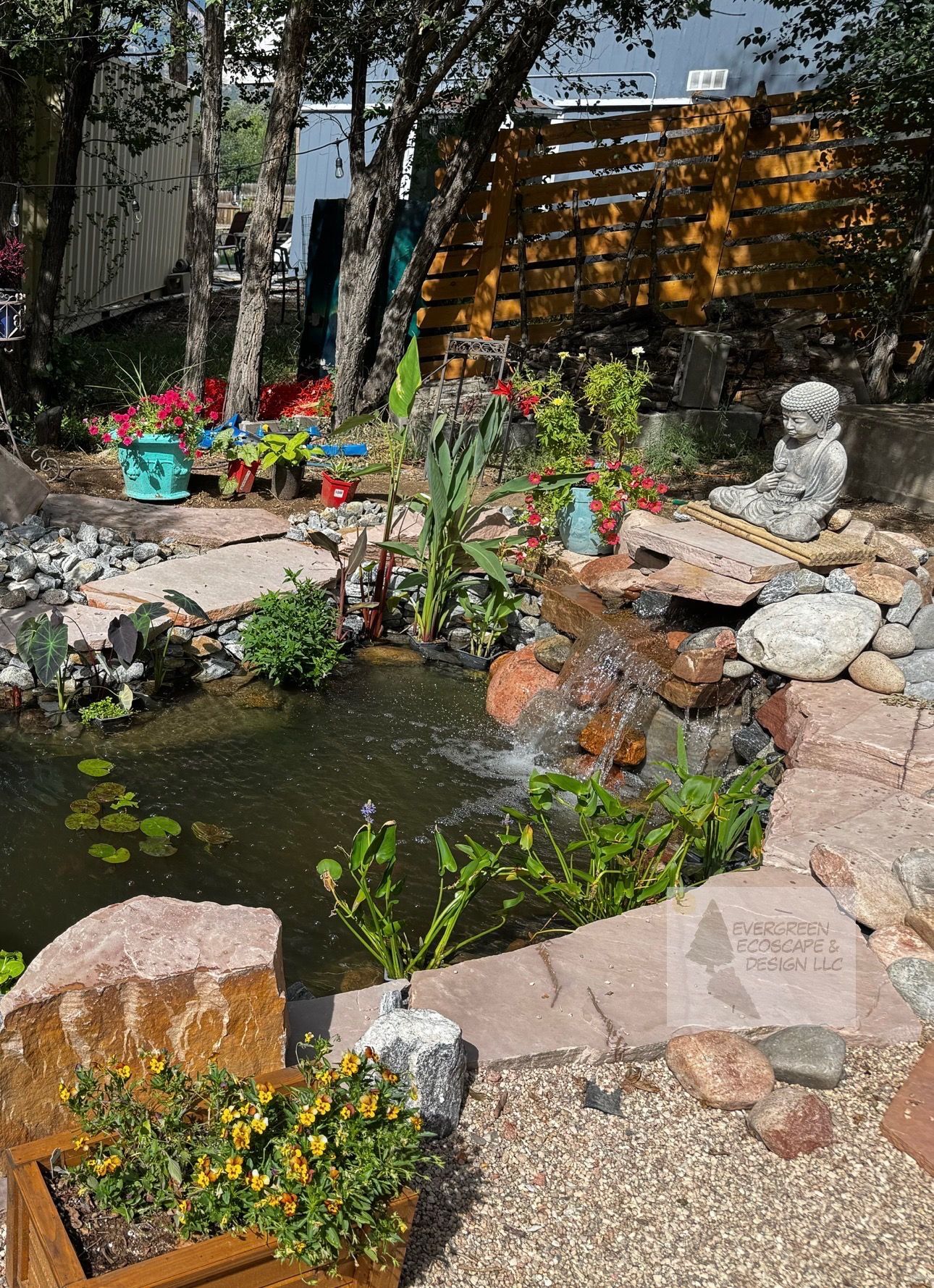 Small garden pond with rocks, yellow flowers, and a stone statue beside trees and a wooden fence.