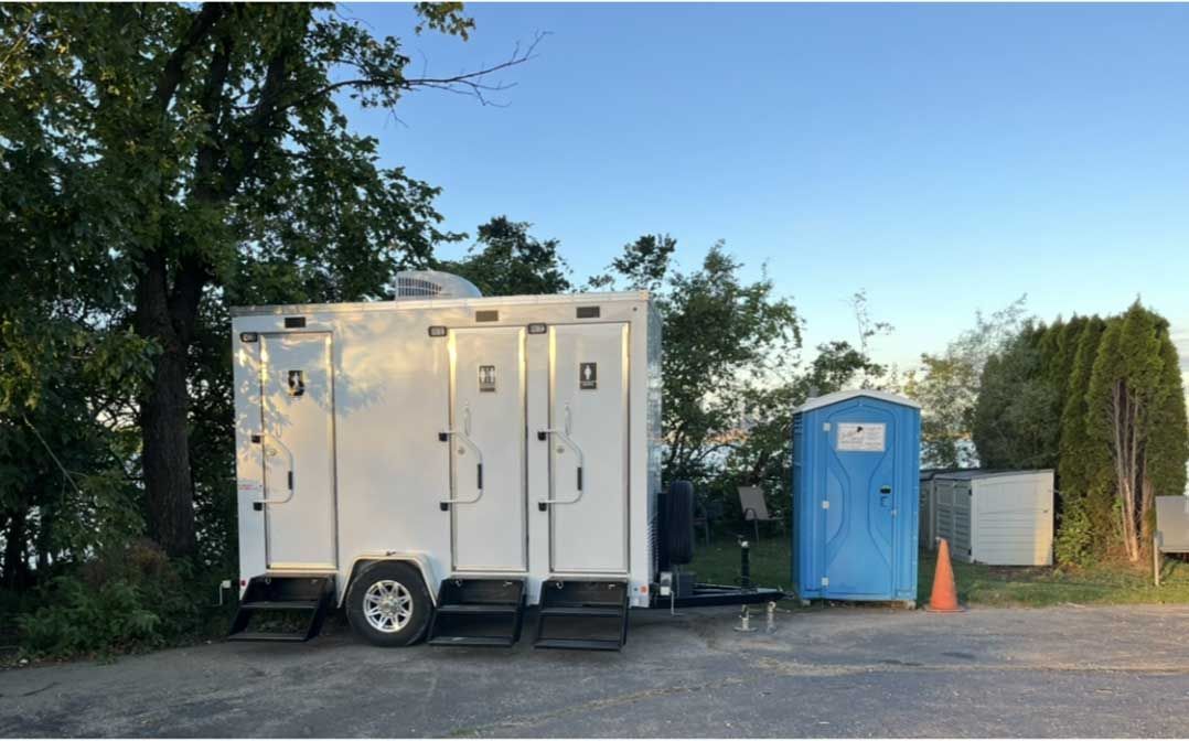 A white portable restroom trailer with three doors parked next to a single blue portable toilet on a gravel lot. A white portable restroom trailer with three doors parked next to a single blue portable toilet on a gravel lot.