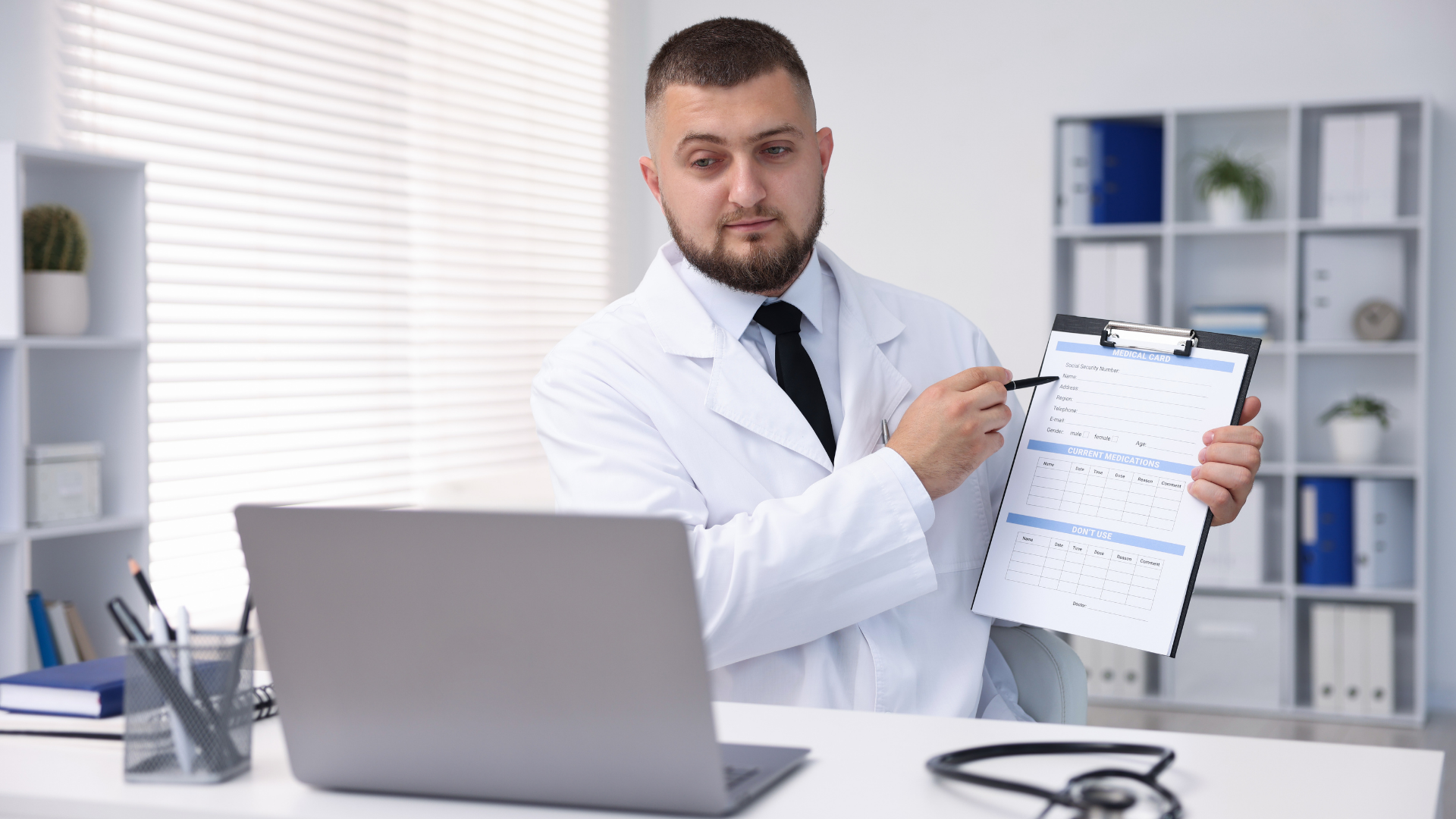 Doctor in white coat holding a clipboard in a bright office beside a laptop and stethoscope.