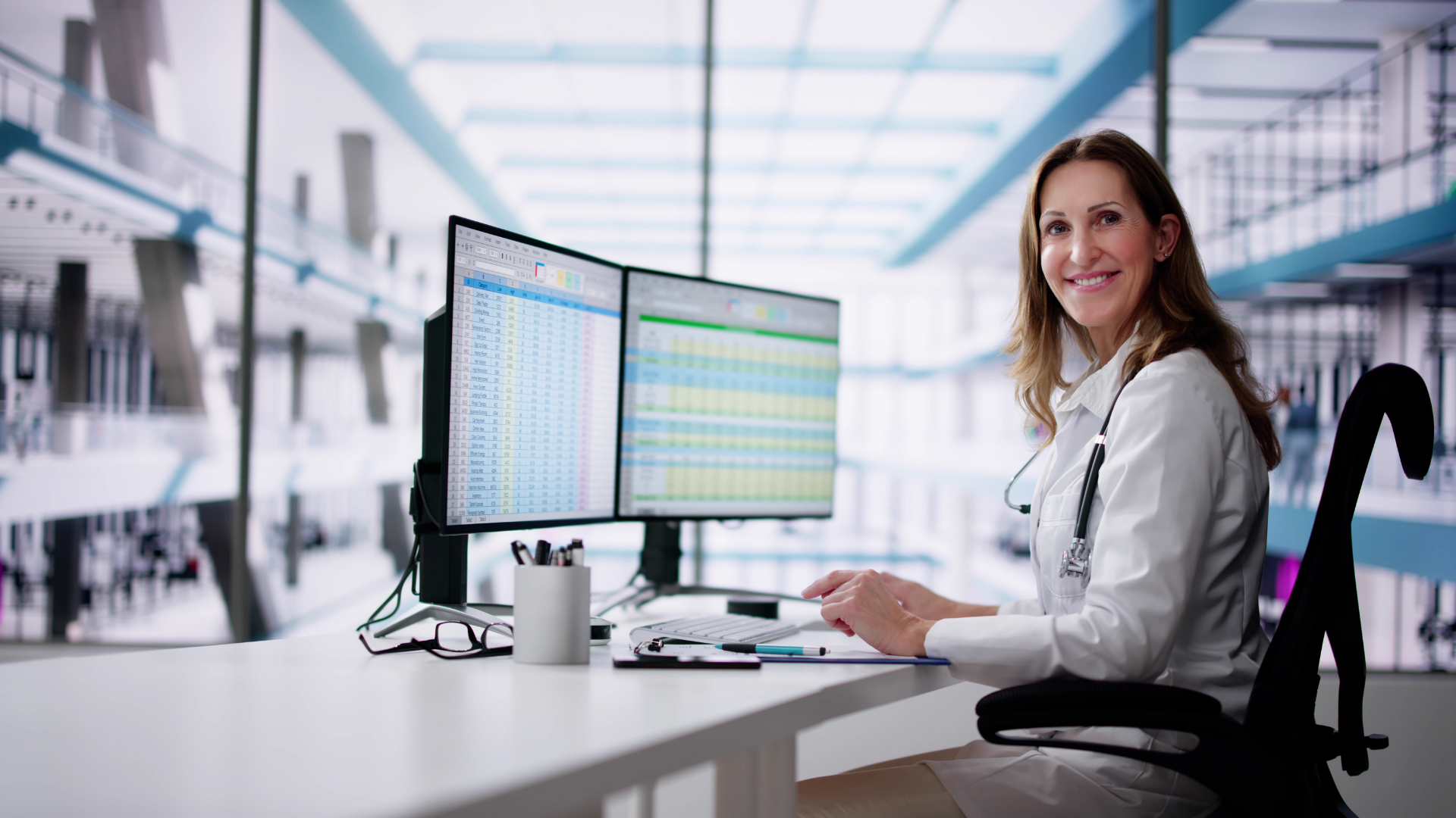 Woman smiling at dual monitors in a bright office, wearing a white coat and typing at a desk