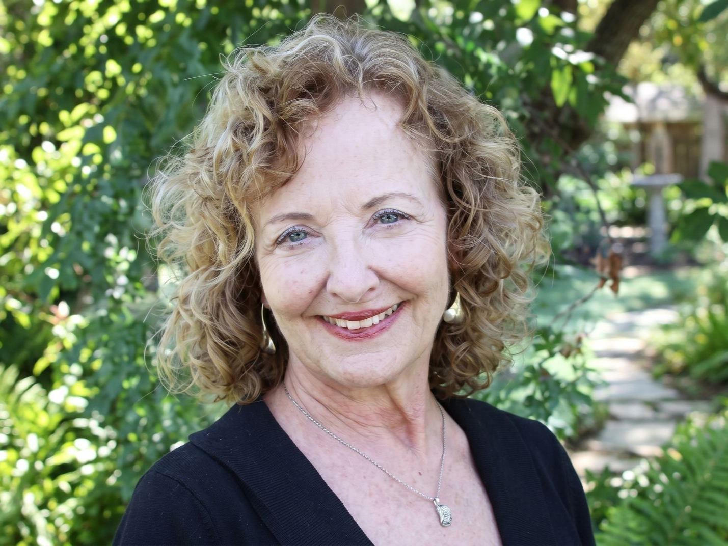 Smiling person outdoors in a garden, with curly hair and a black top.