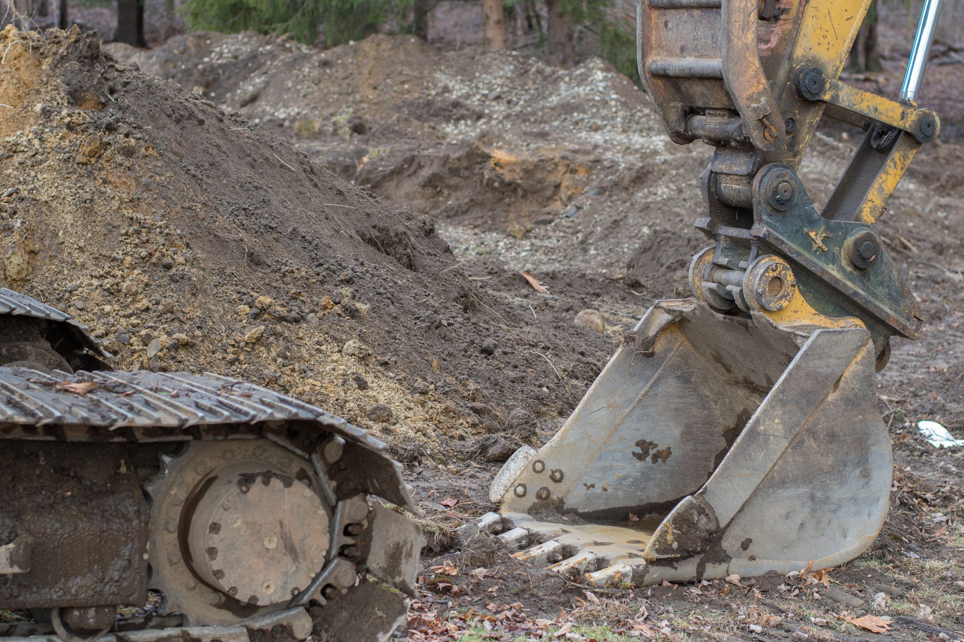 An excavator bucket sits on dirt near its tracks, with a large pile of excavated soil in the background.
