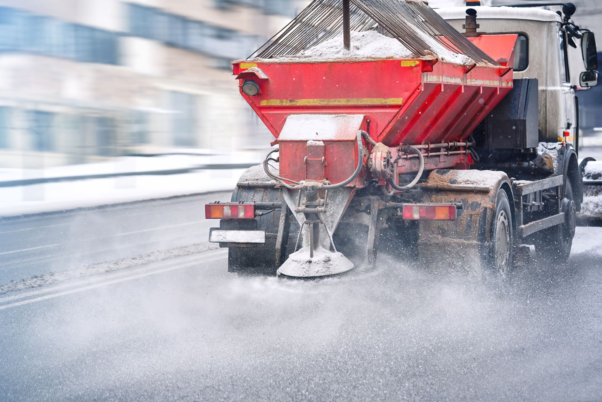 A red salt spreader truck driving on a snowy road, spraying salt onto the pavement.