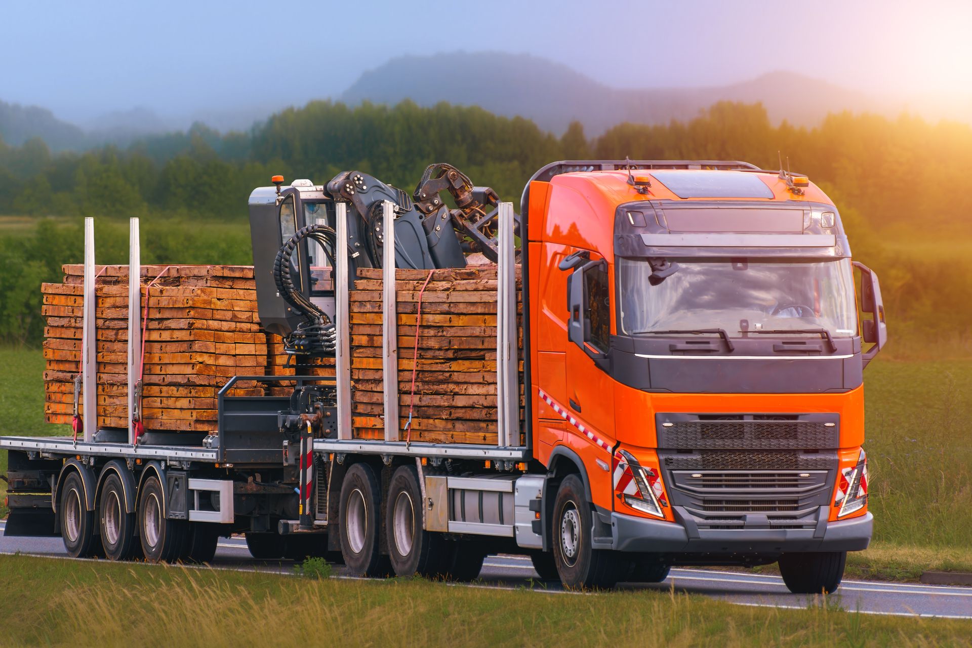 An orange semi-truck carrying a large load of wooden planks on a flatbed trailer, driving on a road at sunset.