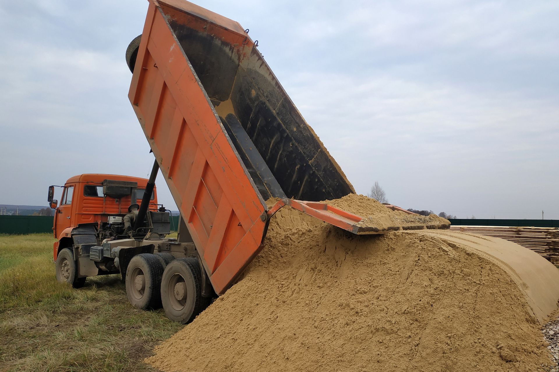 An orange dump truck parked on a field, lifting its bed to unload a large pile of sand.