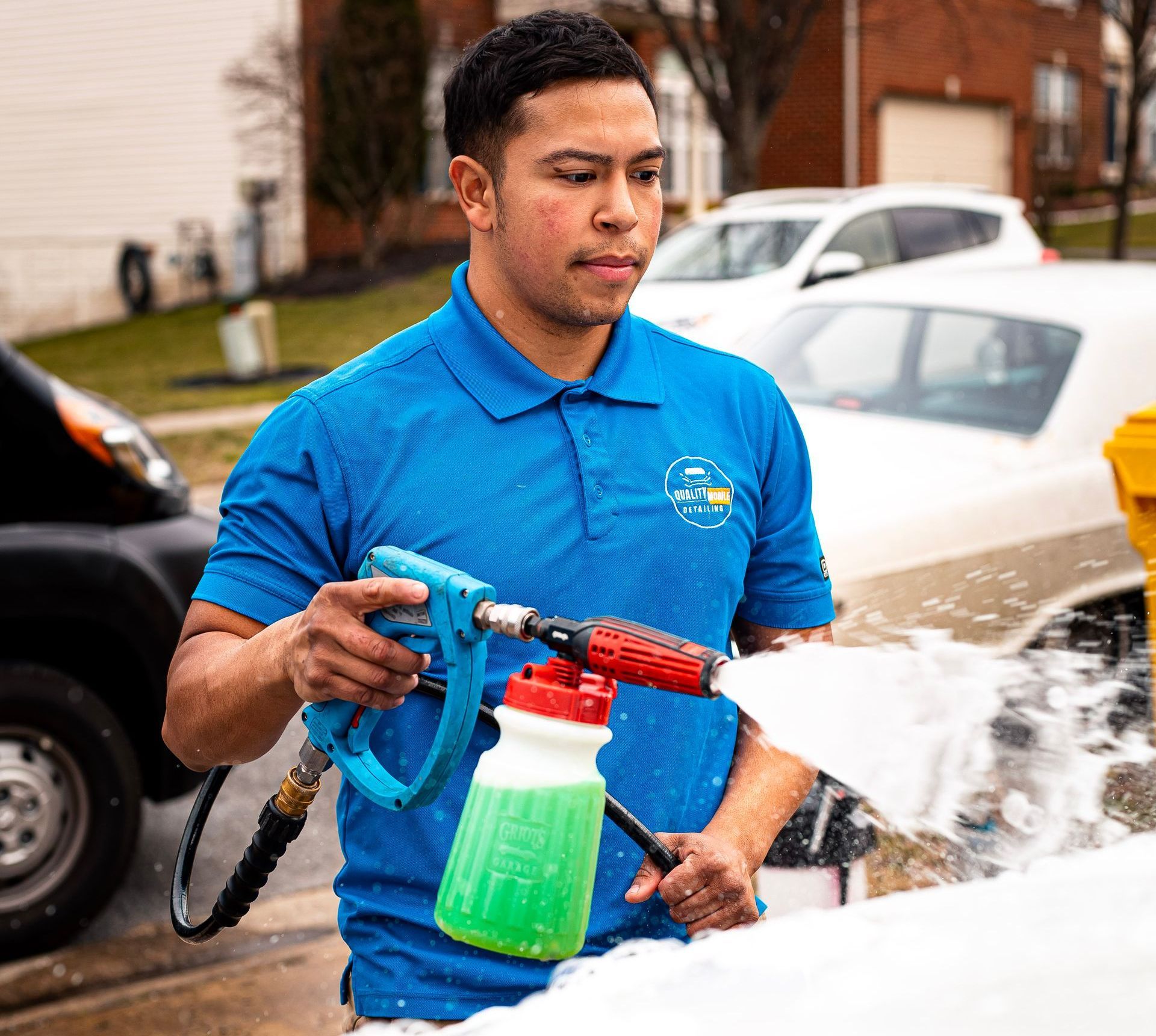 Man using a blue pressure sprayer to wash a car, with water spraying onto the white hood outdoors