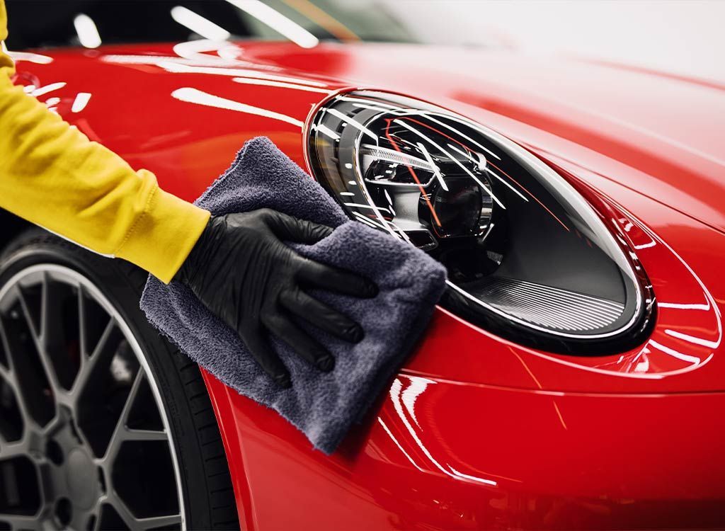 Person in yellow sleeve and black gloves cleaning red car's headlight with a gray cloth.