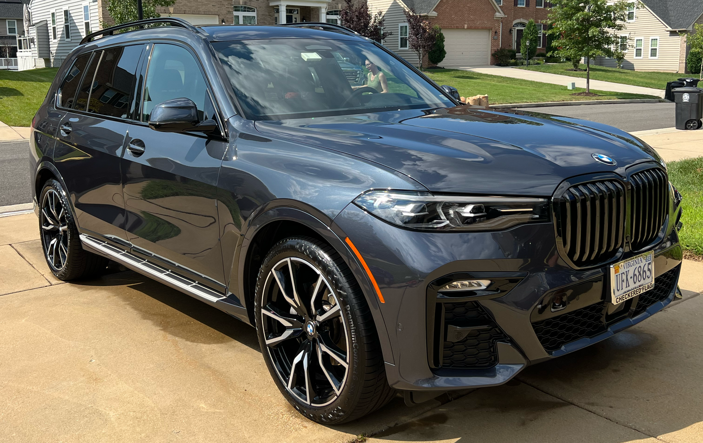 Gray BMW X7 SUV parked on a driveway, black wheels, shiny paint, sunny day.