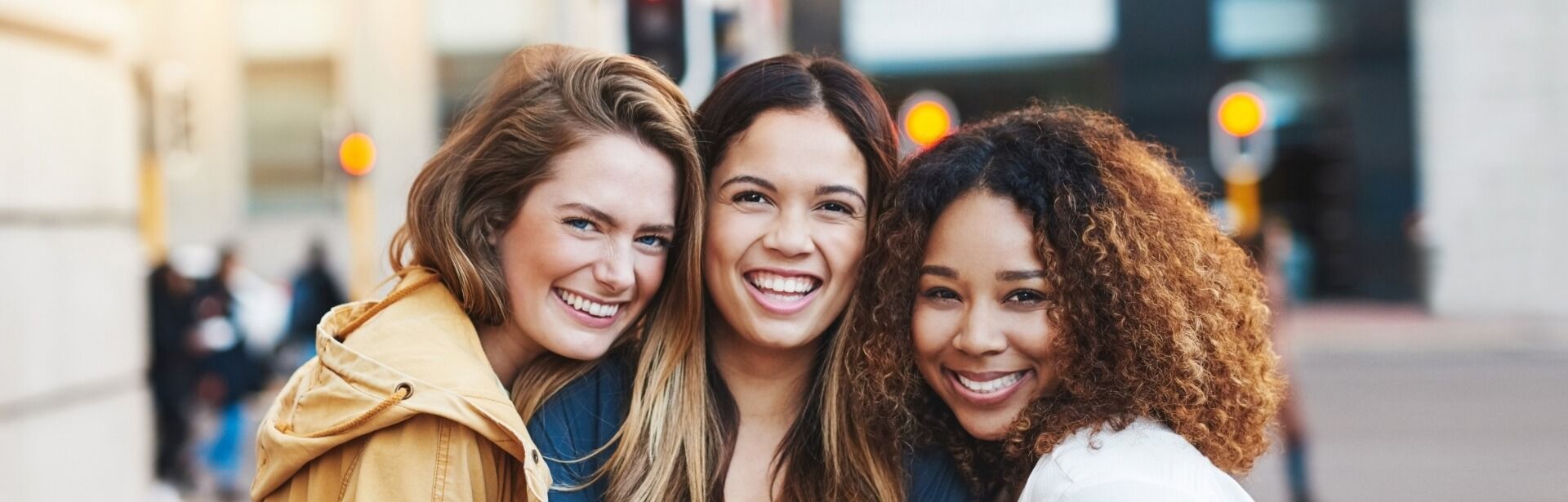 Three people huddled close together, smiling, in an urban setting with blurred street lights in the background.