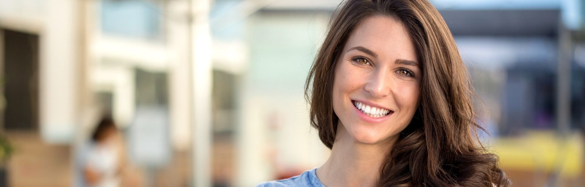 A smiling person with long brown hair looks toward the camera against a blurred outdoor background.