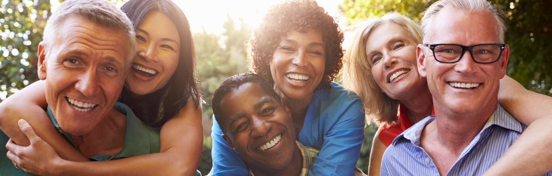 Five smiling adults of various backgrounds are embracing closely together outdoors in warm, natural sunlight.