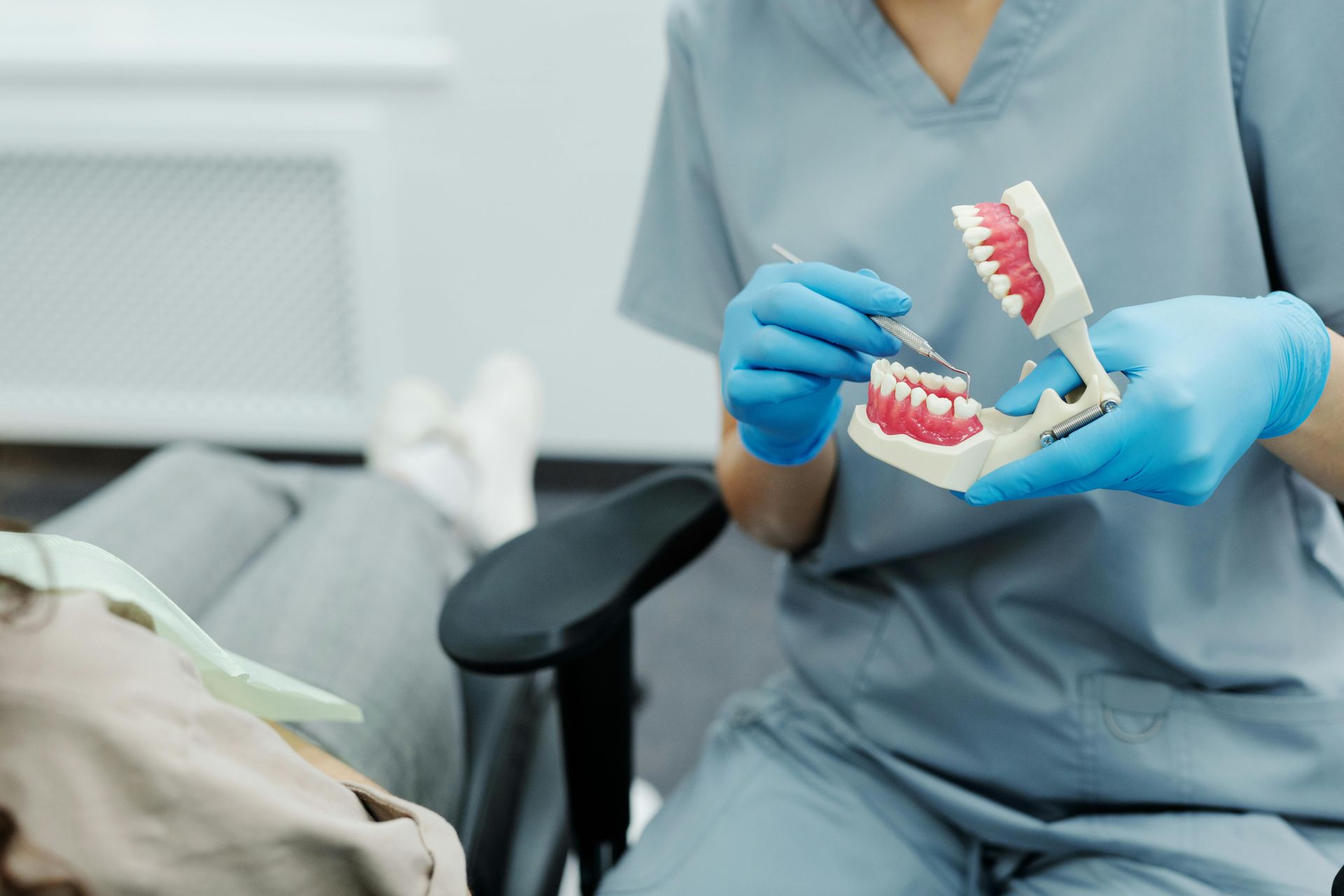 A dentist in blue gloves holds a model of teeth and a dental instrument, explaining a procedure to a patient.