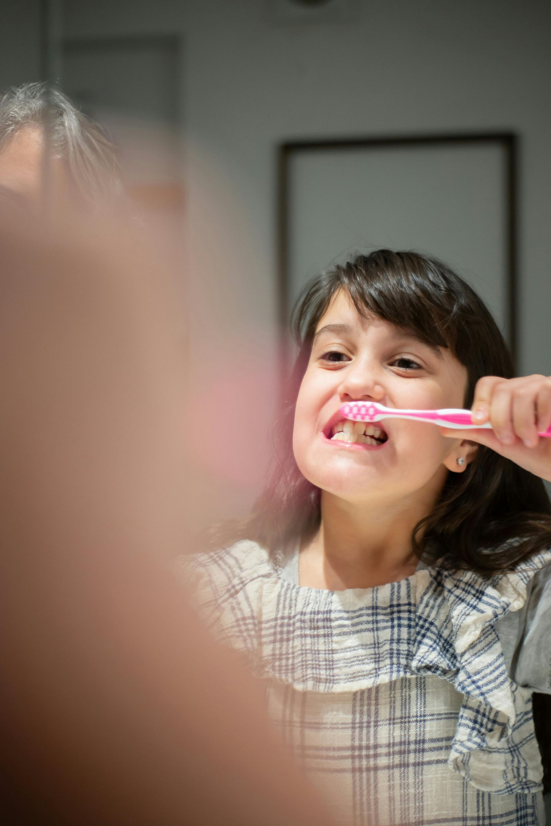 A person brushing their teeth in front of a mirror, wearing a plaid top with ruffled shoulders.