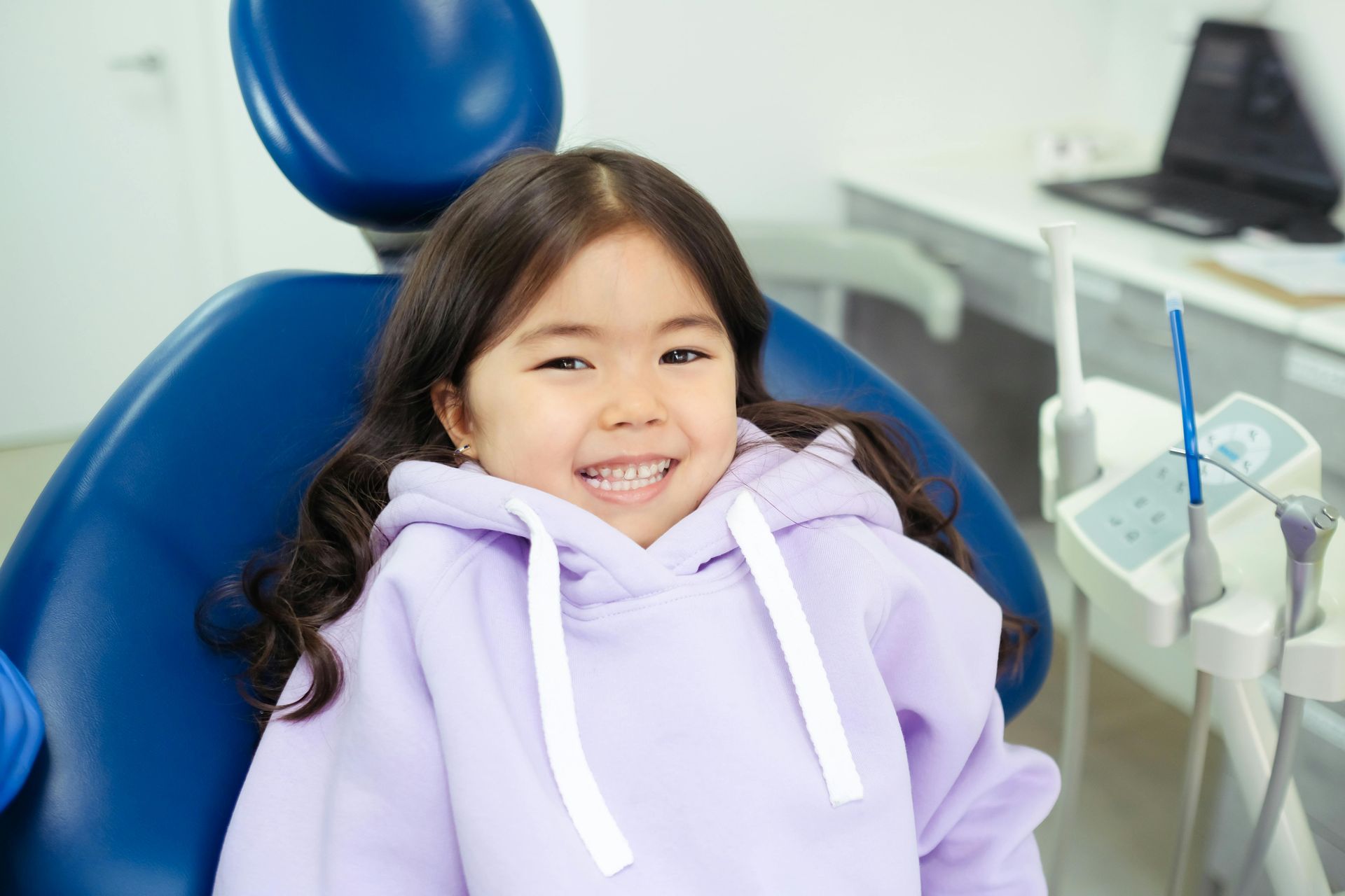 A child smiles while sitting in a blue dental chair in a clinical office setting.