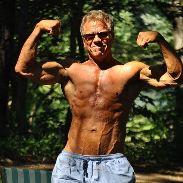 Shirtless man flexing biceps outdoors in sunglasses, standing by a pool in sunlight