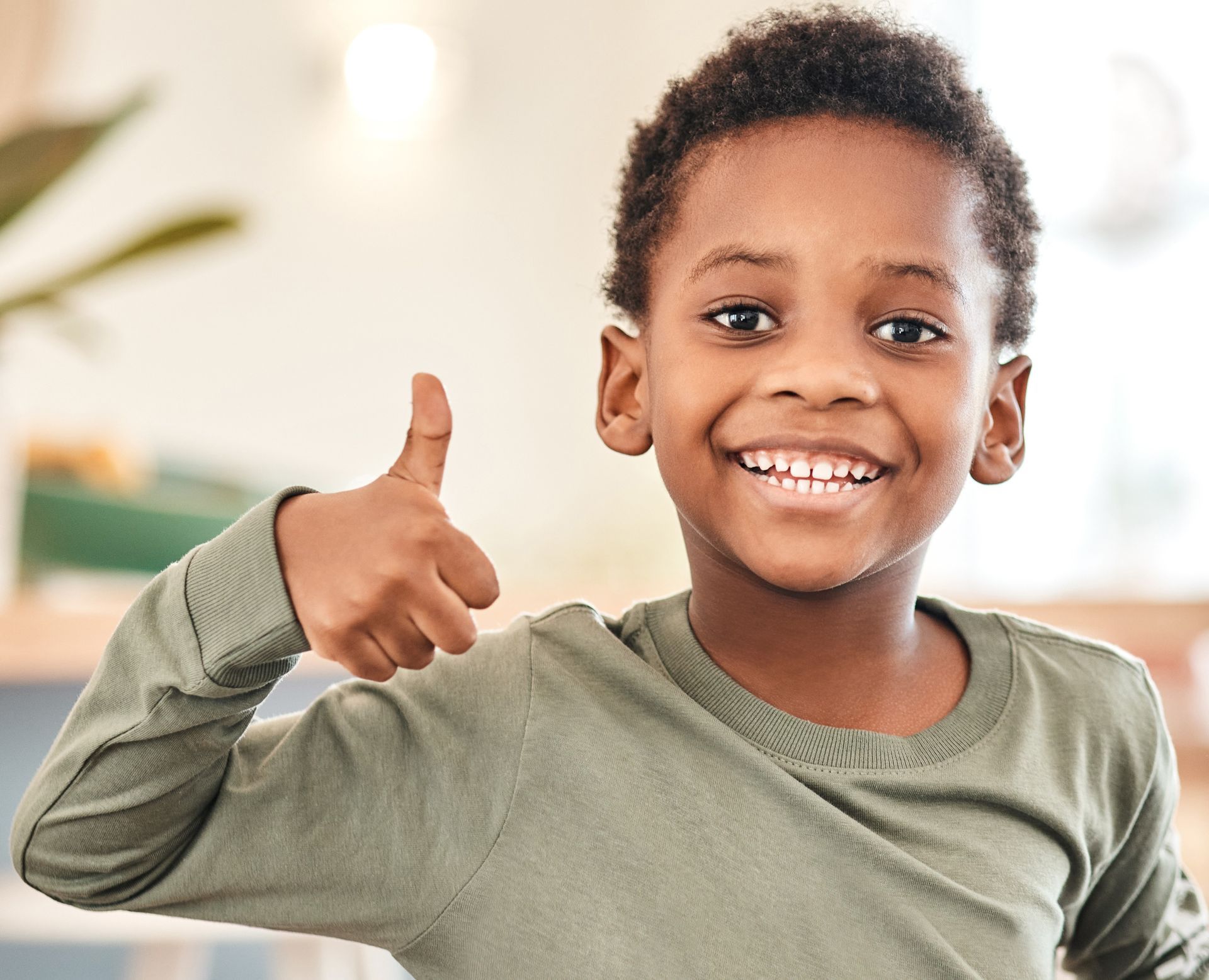 Smiling child in a green shirt giving a thumbs-up indoors