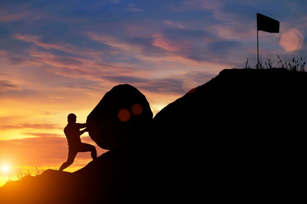 Silhouette of a climber pushing a boulder up a hill at sunset, with a flag on the peak