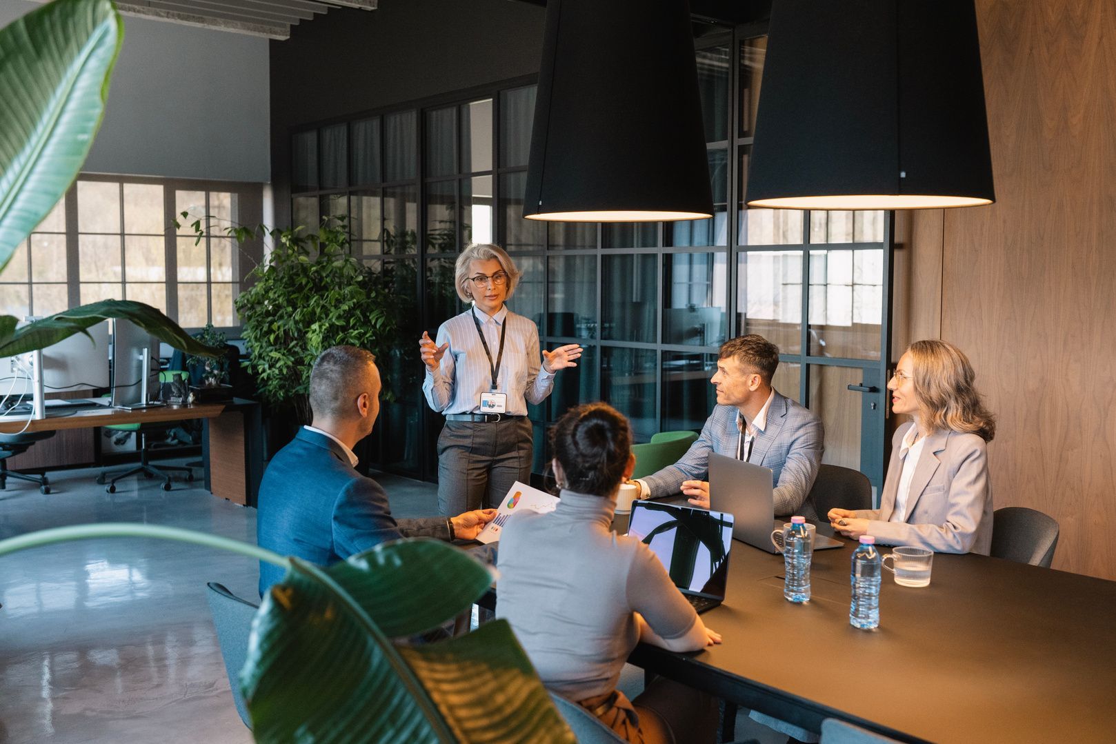 Business meeting in a modern office lounge, with one person presenting to four seated colleagues.