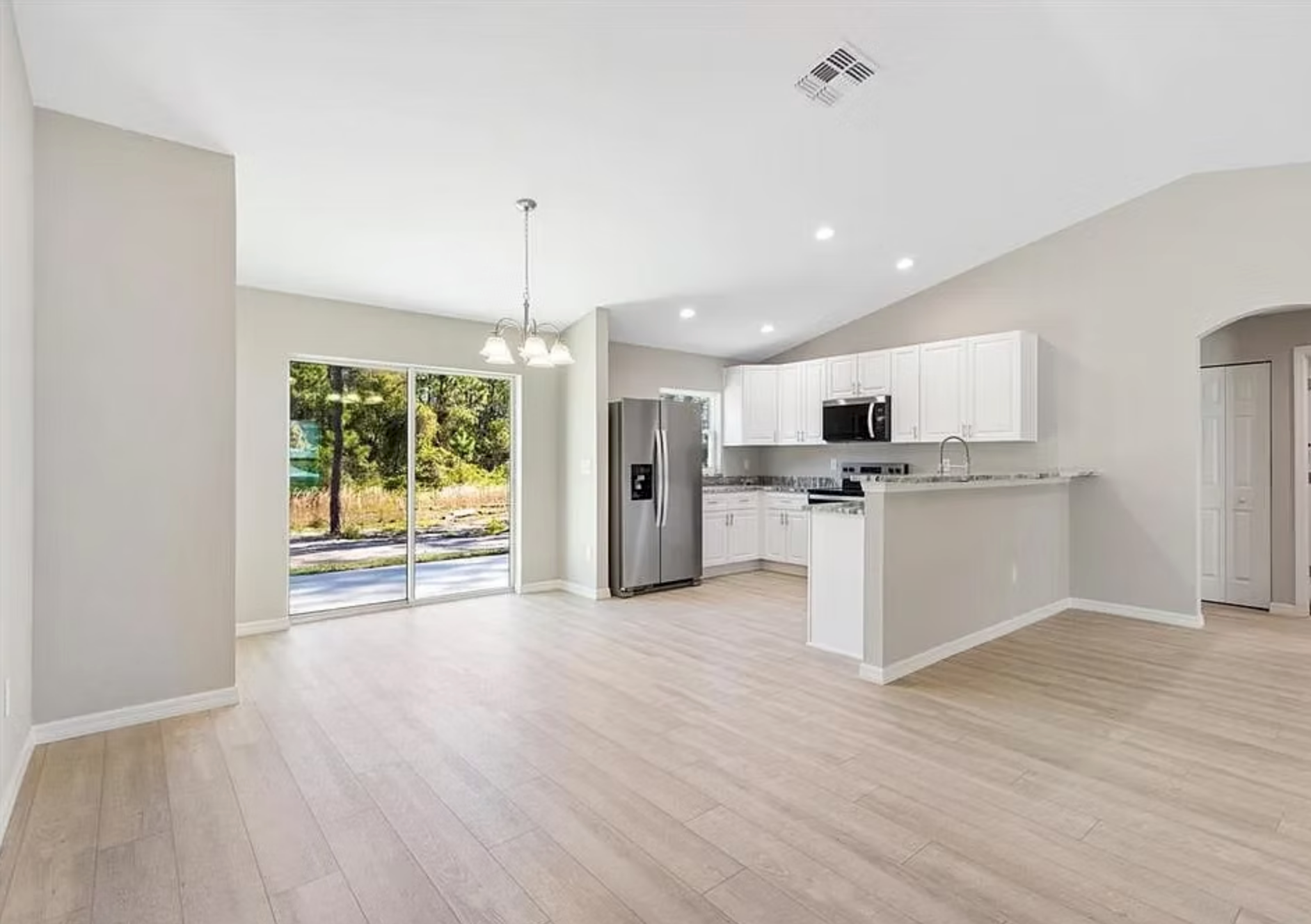 Bright open living room with white kitchen, light wood floors, and sliding glass doors to greenery
